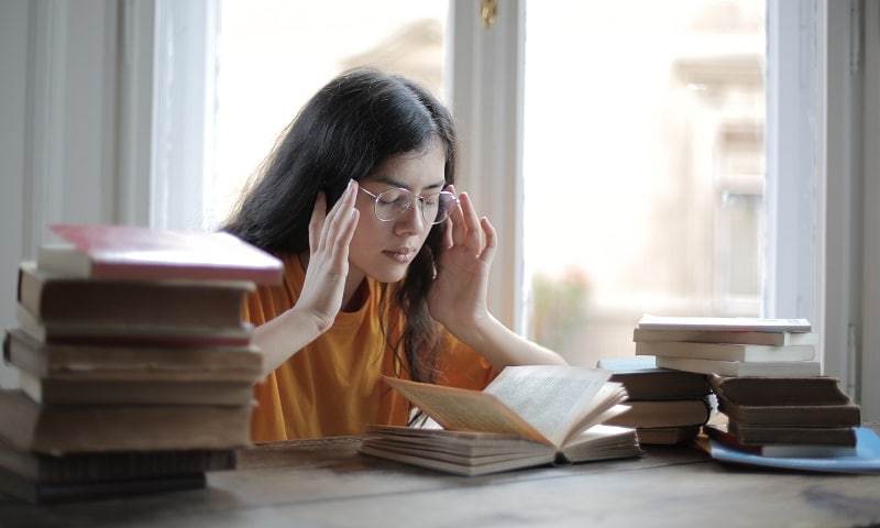 A Girl sitting on a chair wearing eye glasses pressing her head